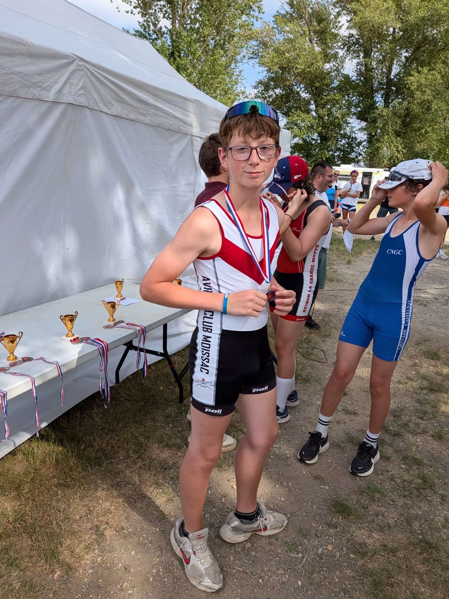 Jeune rameur en maillot blanc et rouge, médaille au cou, devant une table de coupes